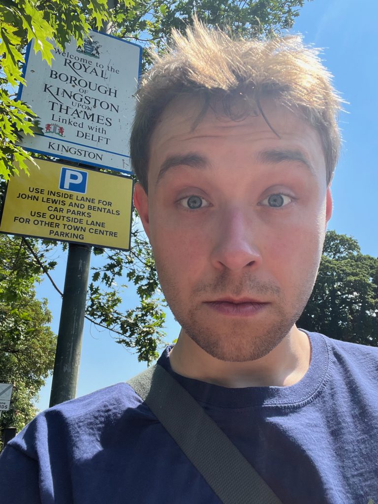 A selfie of the blog's author, looking down at the camera. Behind him is a white rectangular sign with a blue border. At the top of the sign is a coat of arms, then the black text: "Welcome to the ROYAL BOROUGH of KINGSTON upon THAMES". The lower-right strokes of the R in "Royal" and the K in "Kingston" are extended pretentiously below the baseline of the text. Below this is says "Linked with DELFT", with Delft's own coat of arms shown to the left of its name; then, below a blue line, it says "KINGSTON". Beneath this sign is another, a yellow rectangle with a black border. It shows the white-on-blue P road sign for car parks, and below this is black text: "Use inside lane for John Lewis and Bentals [sic] car parks. Use outside lane for other town centre parking". In the background of the photo, behind both, some trees can be seen. The sky is, of course, a cloudless blue.