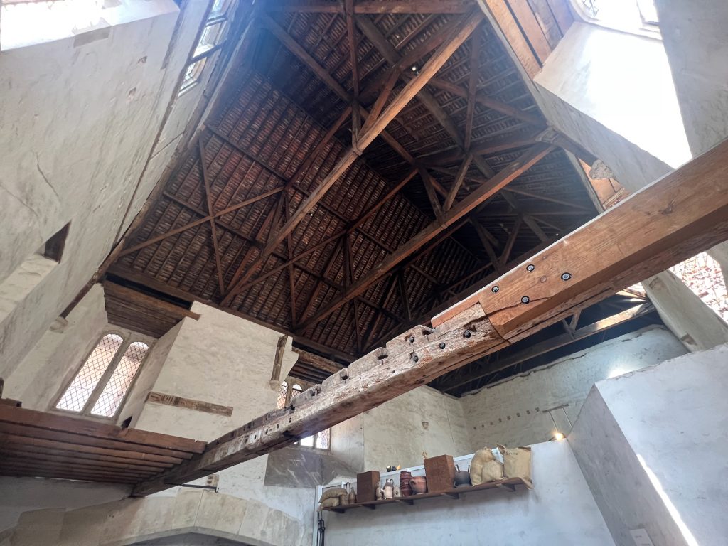 Looking up at an old, high, hipped roof, showing the wooden framework supporting wooden slats. The view is veyr wide-angle, with visible perspective. The roof is above whitewashed walls; a long wooden crossbeam cuts across the picture from the right-hand foreground to the back wall, with shorter beams bridging from the main one to the left-hand wall. At the upper level of the wall there are arched windows with leaded lights. Towards the bottom, the top of a fireplace arch can be seen, along with a half-height wall; on the wall is a wooden shelf, showing historic kitchen supplies.