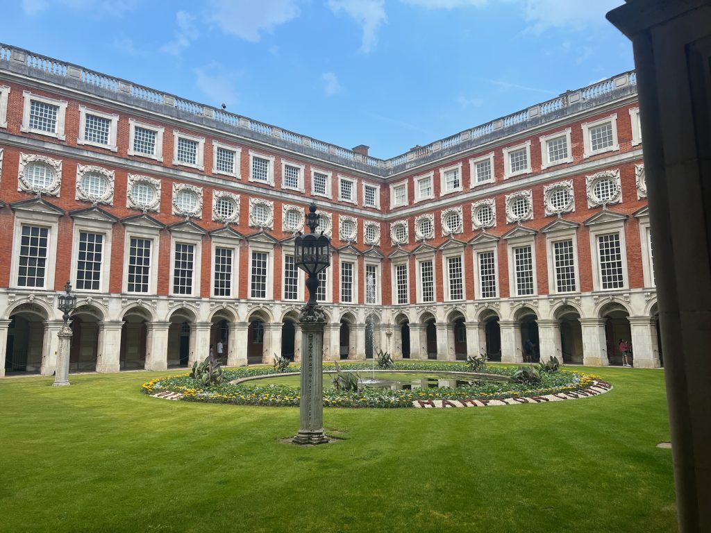 A diagonal view across a rectangular courtyard. Both walls have the same sequence of elements, closely spaced: on the ground floor, an arched colonnade; on the first floor, rectangular divided-light sash windows with a pediment atop each; on the second floor, circular divided-light windows with a decorative stone wreath surrounding them; on the top floor, square divided-light sash windows. The colonnade and window surrounds are of white stone, along with a strip between the second and top floors and a balustrade along the roof; the remainder of the wall is of red brick. The courtyard contains a fountain, which shoots a thin column of water from the middle of a circular pond. The pond is surrounded by planting, which in part spells out words: "2023" on the left and "HAMPTON COURT…" on the right. The rest of the courtyard is a lawn, with four lanterns towards the corners: these are a couple of metres high, and have metal-and-glass lights atop decorative stone columns. The sky is blue.