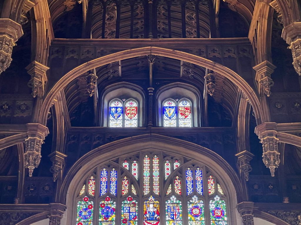 Looking up towards the upper wall of a large, highly ornamented hall. The pointed arches of the vaulted ceiling dominate the top of the picture, showing their gilded detailing. At the top of the wall are two pointed-arched windows, each divided into two smaller ones; the stained-glass windows have as their main element the heraldic shields of France, England, Ireland, and Wales. Below this is a much larger pointed-arched window, divided into smaller panels by stonework; this is again dominated by heraldic elements, but shows the letters "H" and "R" near the middle, below which is a waist-up portrait of Henry VIII.