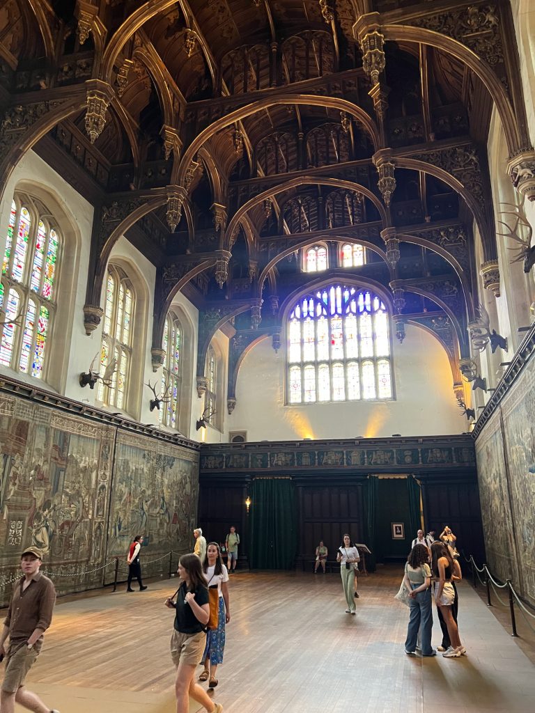 A fairly large, high-ceilinged hall with a vaulted wooden roof. There are staned-glass windows on the upper walls, with decorative tapestries on the lower parts of the left- and right-hand walls; the rear wall is panneled, with two large doorways in it. Tourists mill around on the wooden floor.