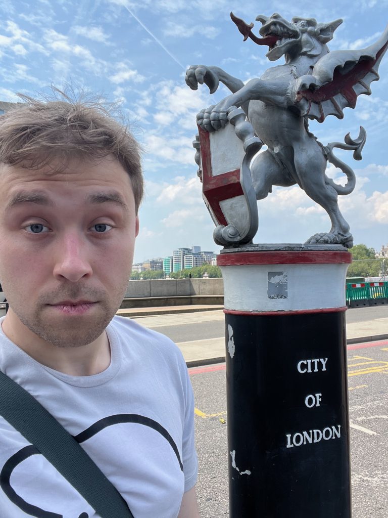 A selfie of the blog’s author, looking half-asleep and wearing a grey Herdy t-shirt. He stands next to a black-and-white post with red detailing, above which is an iron statue of a dragon, painted silver, with red details on the underside of its wing and a red tongue. The dragon stands on its hind legs, with one front talon aloft, and the other holding the top of a shield, which shows the City's coat of arms (a St George's cross, with a red sword in the top-left quadrant). Behind both human and dragon is the road, which is about to cross the Thames on a bridge; the river isn't visible, but some high-rise buildings are on the far embankment. Surprisingly, there are fluffy white clouds across the blue sky.