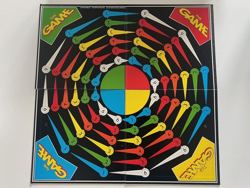 A game board. The playing area has spaces arranged in six concentric circles, each of which has ten spaces around the circle (so there are ten spokes). The spaces are numbered along each spoke from 1 in the middle to 6 at the outside, or vice versa. Each set of numbered spaces is the same colour (blue for 1, green for 2, yellow for 3, orange for 4, red for 5, and white for 6). In the centre of the centre circle is a disc, divided into four quarters coloured green, red, yellow and blue. The corners of the board all contain the game logo, which says "THE GAME" in writing with a perspective effect. The background colour of each corner matches the quarter-circle nearest it in the centre.