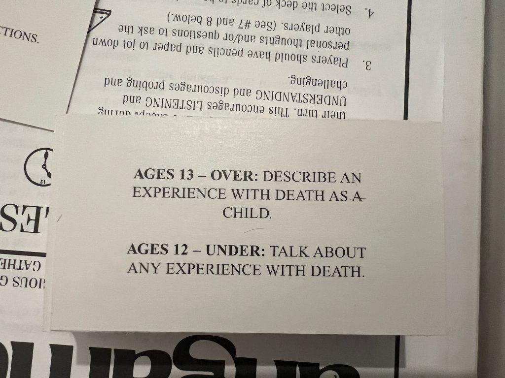 A board game card that says "Ages 13 – over: describe an experience with death as a child. Ages 12 – under: talk about any experience with death." It is placed on top of the rules for the game. The visible part, which is upside-down, says "… during their turn. This encourages LISTENING and UNDERSTANDING and discourages probing and challenging. "3. Players should have their pencils and paper to jot down personal thoughts and/or questions to ask other players. (See #7 and 8 below.) "4. Select the deck of cards…"