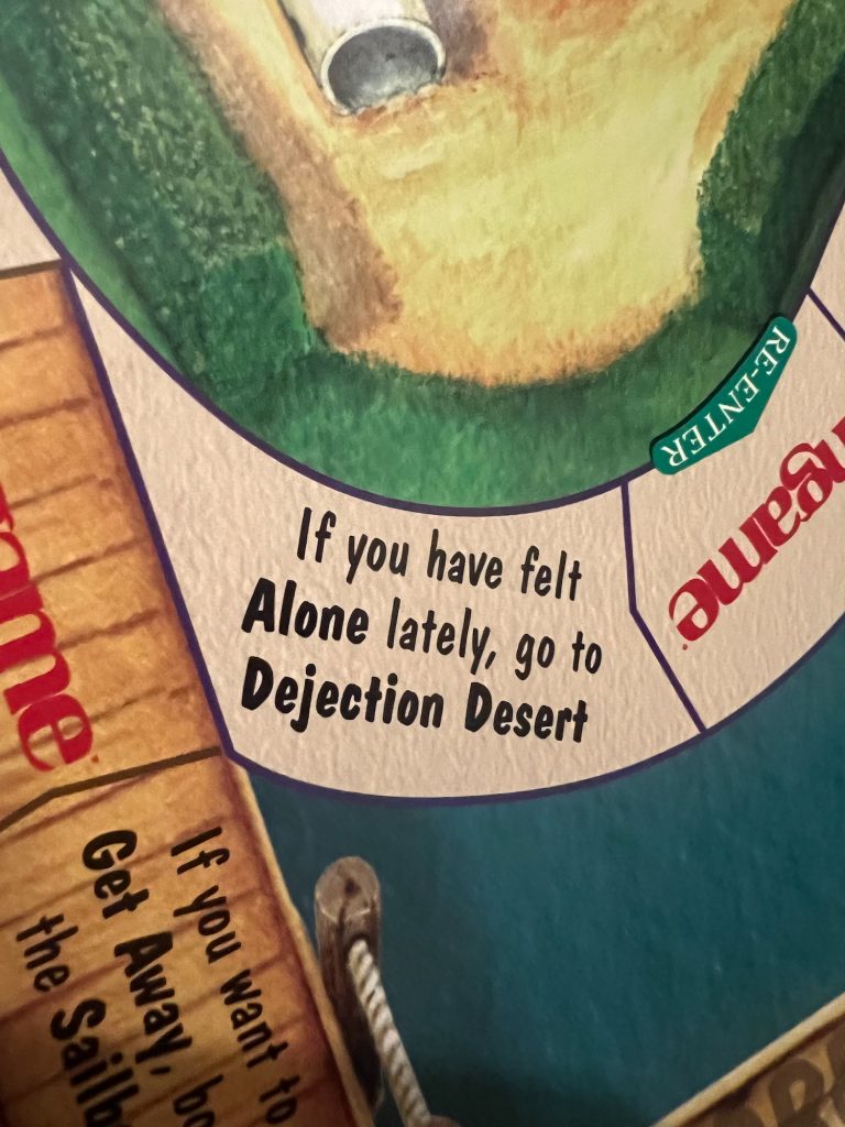 A close-up of a space on a board for a board game, which reads "If you have felt Alone lately, go to Dejection Desert". The words "Alone" and "Dejection Desert" are in bold. The path is beige, curving around between two spaces with "ungame" printed on them. The left-hand space and another space are decorated like decking, with text on the second that says "If you want to Get Away, b[oard] the Sailb[oat]". Above the curve of the main track is an illustration of what looks like a sandpit with a plastic tunnel, seen top-down, with an arrowhead marked "RE-ENTER" pointing back onto the path. Below the path is a sea-blue colour.
