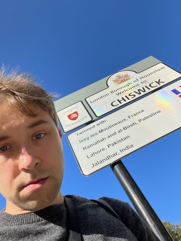 The blog's author, standing next to two rectangular signs that share two black poles. The top one is a grey board with square corners, and two signs on it. On the left is a small rectangular one (white with a rounded black border) that shows a red shield, depicting three swords beneath a crown. Arching above the shield are the words "County of", and mirroring the curve below the shield is the word "Middlesex". The right-hand sign is rectangular (again, white with a rounded black border), but with a hump protruding from the middle of the upper edge. Within this hump is the borough coat of arms, and in the rectangular part are the words "London Borough of Hounslow, Welcome to CHISWICK". The lower sign has rounded corners, and is white with a black border. The text says "Twinned with: Issy-les-Moulineaux, France, Ramallah and al-Bireh, Palestine; Lahore, Pakistan; Jalandhar, India". Aligned with each twin settlement is the corresponding flag, although most are cropped off, with the only visible parts being some of the French flag and a tiny corner of the Palestinian flag. The overall view is looking up, so the background is a solid blue sky much like the previous day of doing this pointless activity, with a small section of three visible at the bottom-right corner behind one of the black poles.