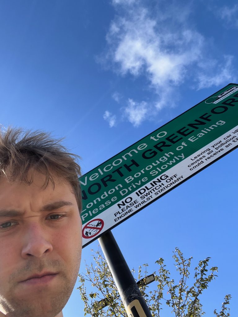 The blog's author in front of a sign; this time you can only see his head, the upward angle is so steep. Behind him is a rectangular sign with a black border; the top two-thirds or so of the background is green, the bottom third being white. On the green section, it says "Welcome to" in white text, then "NORTH GREENFORD" in black, and then, in white again, "London Borough of Ealing, Please Drive Slowly". At the top-right is the Ealing council logo: a tree, next to the word "Ealing" in white on a black box, and the council's web address in a white box beneath that. On the white section is a pictogram, showing a car spewing fumes at an adult and a child, all crossed through in red, inside a red circle. To the right of this it says: "NO IDLING: Please switch off engine while stationary". The text to the right of this is partly cut off, but what can be read says "Leaving your car idl… causes air pollution… could receive an £…". A small amount of foliage can be seen behind one of the black poles. The sky is blue with some fluffy clouds.