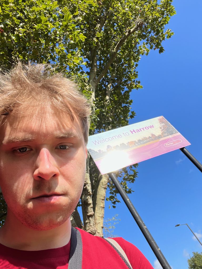 The blog's author standing by a road, wearing a red t-shirt. Behind him are some trees, and a rectangular sign supported by two black poles. The top part of the sign shows a photograph of a green space surrounded by houses; printed over the sky are the words "Welcome to Harrow" in purple. The sign below the photo is purple, although part of this to the left is obscured by the reflection of the sun. At the right of the purple part is the council logo, which says "Harrow Council" inside an oval. Little else but blue sky can be seen in the photo, except for a lamppost and another tree in the distance to the right.
