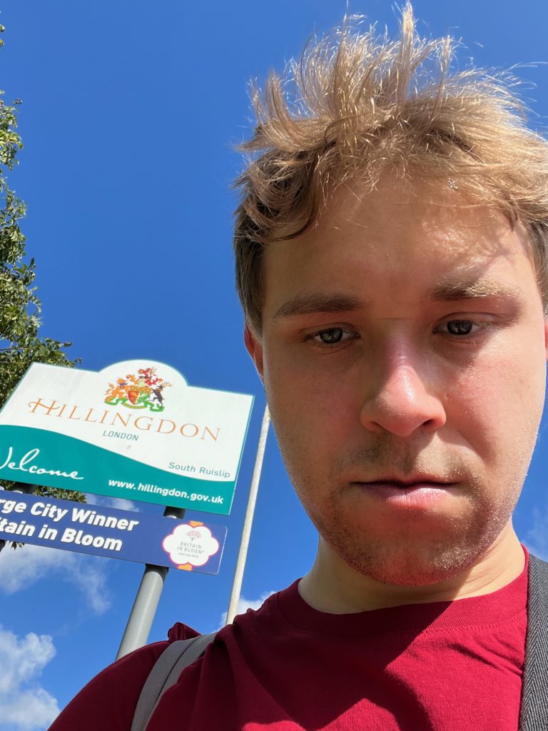 The blog's author looming over the camera, hair flying in the wind. Two signs are visible behind him over his right shoulder, one above the other on the same grey poles. Both are rectangular, except that a hump protrudes from the middle of the top sign. The top sign is divided along a swooshing line into a white upper section and a turquoise lower one. In the white part, a coat of arms is under the hump at the top, with yellow-orange writing underneath that says "Hillingdon". The crossbar of the "H" is a similar swoosh shape to the divider on the sign. Below that, much smaller, it says "London" in turquoise, and then "South Ruislip" appears at the bottom-right within an upward-curling part of the swoosh. On the turquoise part is "Welcome", in a script font, and the council's web address. The second sign, below, is dark blue, and says in white "[La]rge City Winner, [Bri]tain in Bloom", with the Britain in Bloom logo shown on a white flower shape at the right. Nothing is visible in the background except part of a tree and part of a lamppost, and the rest is blue sky with some clouds lower down.