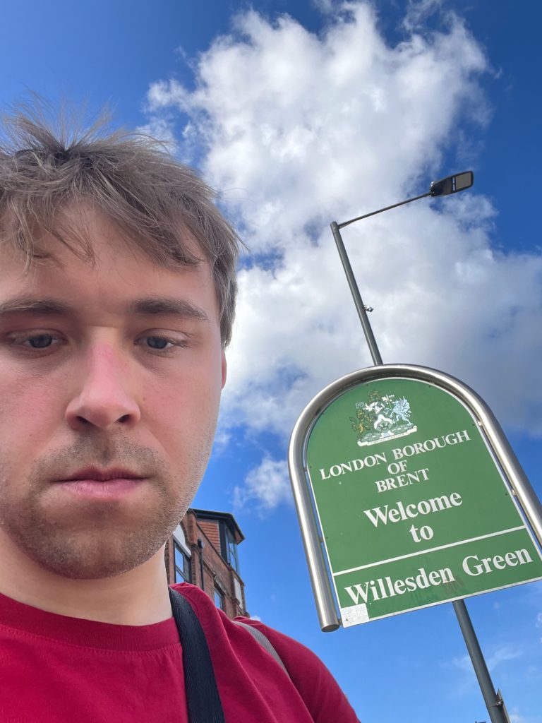 A selfie of the blog's author, wearing a red t-shirt and staring impassively at the camera; he takes up much of the left-hand side of the frame. Behind him is a green sign, supported by a silver pole that rises to the right of the sign, arches over the sign in a semi-circle, and finishes well above the ground at the bottom-left corner of the sign. The sign itself is green with white text, and matches the shape of the pole. At the top of the sign is a coat of arms, and below it the text "London Borough of Brent, Welcome to". There is then a horizontal white line running across the sign, with the words "Willesden Green" below that. Behind the sign is a lamppost, and behind the blog’s author can be seen a small portion of a brick building. The sky is partly cloudy.