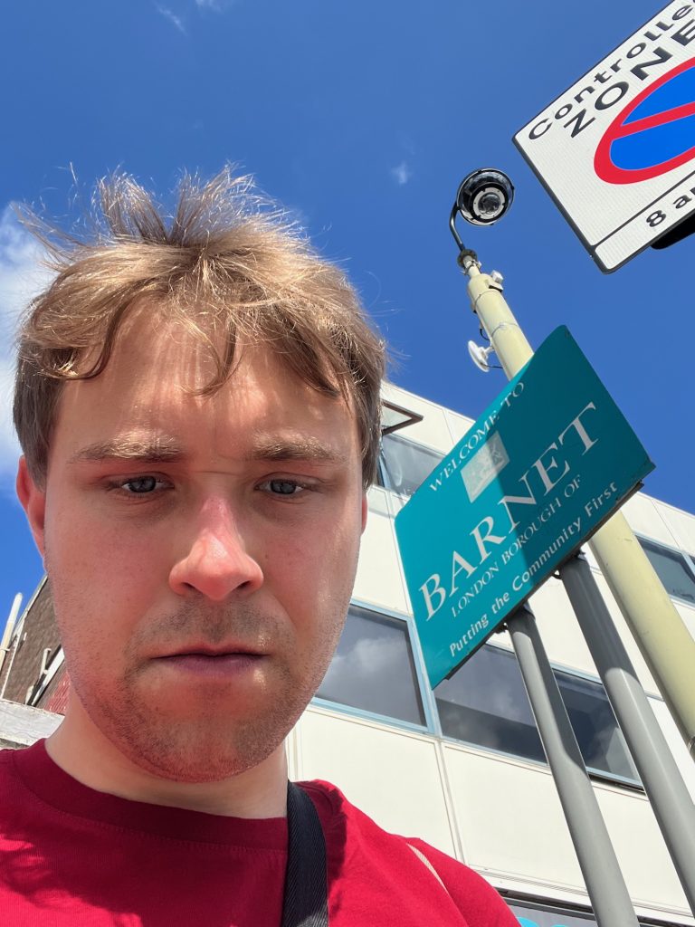 A selfie of the blog's author, who stoops over the camera. To the right of his head, behind him, is a turquoise rectangular sign with white writing, on two closely-spaced grey poles. The first line of the sign says "Welcome to", and then there is a faded image of a coat of arms. Below that the text says "BARNET, London Borough of", and then, after a gap, "Putting the Community First". Other things in the background include what looks like a CCTV camera on a yellow-ish pole, and a controlled parking zone road sign (which starts at 8am, but the end time is obscured). Behind all of those are a mid-twentieth-century office buildign, with a fascia divided into square windows and rectangular plastic panels. The sky is mostly blue.