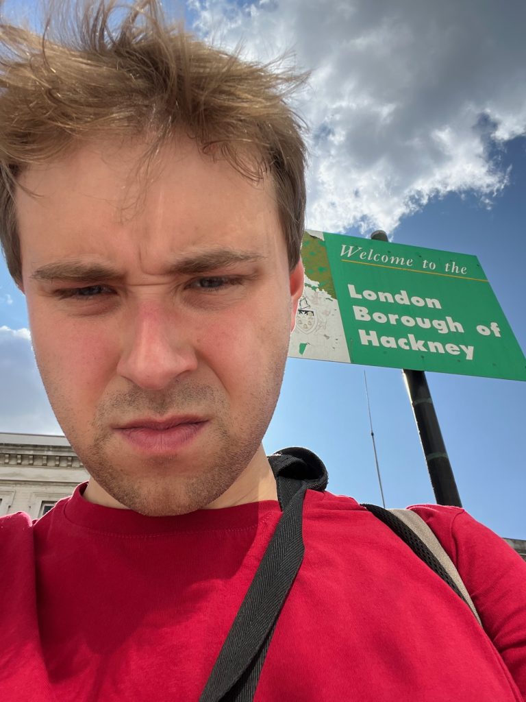 A selfie of the blog's author, taking up much of the frame. Above his right shoulder (the viewer's left) the top of a white-painted building can just be seen, with a decorative freize along the roofline. Above his right shoulder is a rectangular sign on a black pole. The left-hand third of the sign is white, but it appears to be peeling off, and shows a faded image of a coat of arms. The right-hand third is green, with white writing saying "Welcome to the London Borough of Hackney". There is a thin yellow line below the first three words. The sky is partly cloudy.