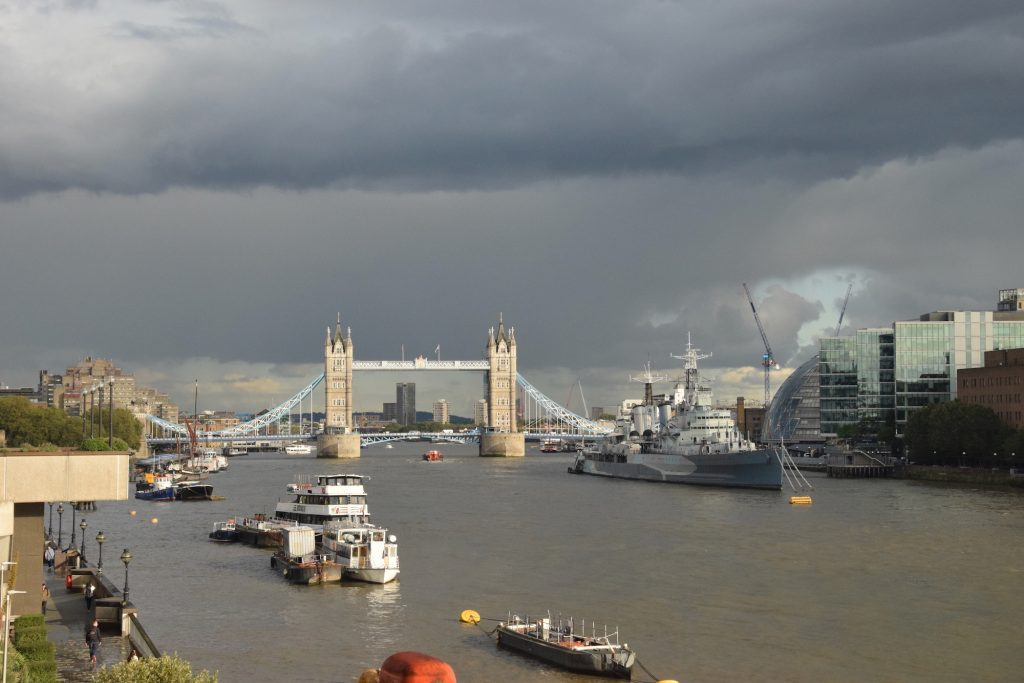 Tower Bridge in London, seen from somehwat upriver (on London Bridge, though London Bridge isn't visible). In front of it, and to the right, is HMS Belfast, and there are other boats on the river. Some glass buildings, include the old London City Hall, line the right-hand bank, while the buildings on the left are predominantly concrete. The river is a murky brown, beneath a sky with almost complete cloud cover; a small patch of blue persists on the right of the image.