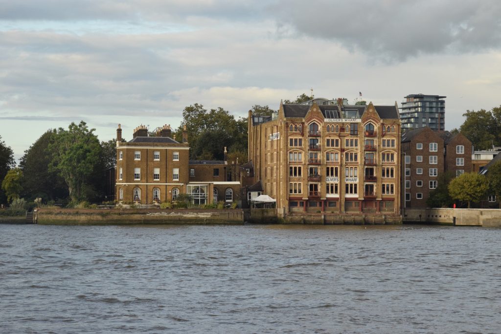 The bank of a river, viewed from the opposite bank. The bank is straight on to the camera. To the left is a garden, with trees visible; next to it is a three-storey yellow-brick house, four window-bays across, with arched sash windows on the ground floor and rectangular ones above. To the right of that, in turn, is a six-storey building made of yellow brick, and six window-bays across. There are three painted signs on the walls of the building which identify it as "Oliver's Wharf". The ground floor has very small windows, and looks like it was once used for landing boats. Above that, the second and fifth columns of windows are slightly recessed, and have red-painted Juliet balconies. There is then a more plain five-storey brick building to the right, slightly recessed. A modern high-rise building rises in the background. The river is choppy and grey; the sky is mostly cloudy.