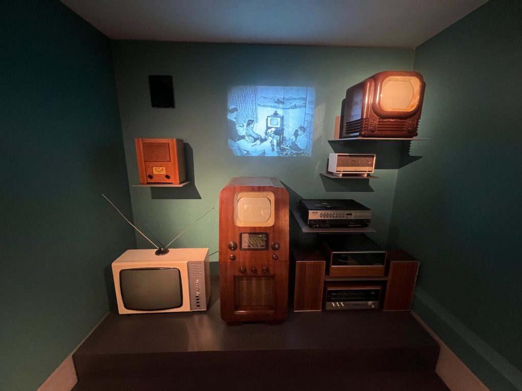 A collection of vintage home entertainment devices in a museum. On a black plinth on the ground are: a white television with two aerials sticking out of the top symmetrically, and buttons and a speaker on a silver recessed panel to the right of the screen; a teleivision with a small screen in a fairly tall wooden cabinet, with knobs, a tuning pointer, and a grille all below the screen; and a wood-and-metal hifi system, with a turntable and main control unit flanked by two speakers. On separate wall shelves are: a wooden radio with a small tuning dial and two knobs; a black-and-silver record player; a white plastic clock radio; and a small wooden television. The walls are painted dark teal, and projected onto the back one is an old black-and-white photograph of a family sitting around a small television. There is also a black speaker on the back wall, though this doesn't seem to be an exhibit.