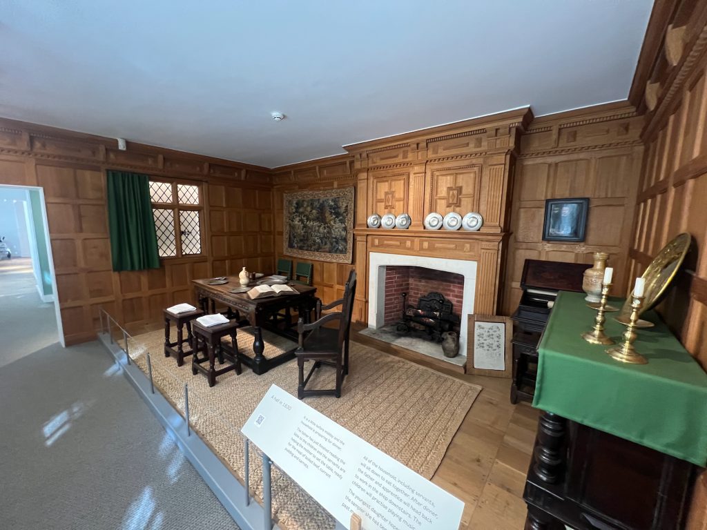 A room decorated as it might have been in 1630. There is wood panelling on all the visible walls, and a window on the left wall in six panels with diamond-shaped leading on the glass, partially obscured by a plain green curtain. On the back wall are hung a tapestry to the left of the fireplace, and a painting to the right; the fireplace itself is lined with brick, and there are sick plates on the mantlepiece. In front and to the left of the fire is a dark wooden dining table and chairs, with a book and some metal plates arranged on it. To the right of the fireplace are a print in a plain wooden frame (leaning against the mantlepiece) and two wooden cabinets. The one against the side wall has a green cloth over its top, on which are placed two yellow-metal candlesticks, a yellow-metal plate, and a pottery jar. The floor is of bare floorboards, covered under the dining table by a straw-like woven mat. The foreground reveals that this is a museum: the room is separated from a pathway by a wire held by plain metal poles, and there is a sign describing the room. In front of the poles is plain carpet, leading to a plain doorframe cut through the panelling which leads through the museum.