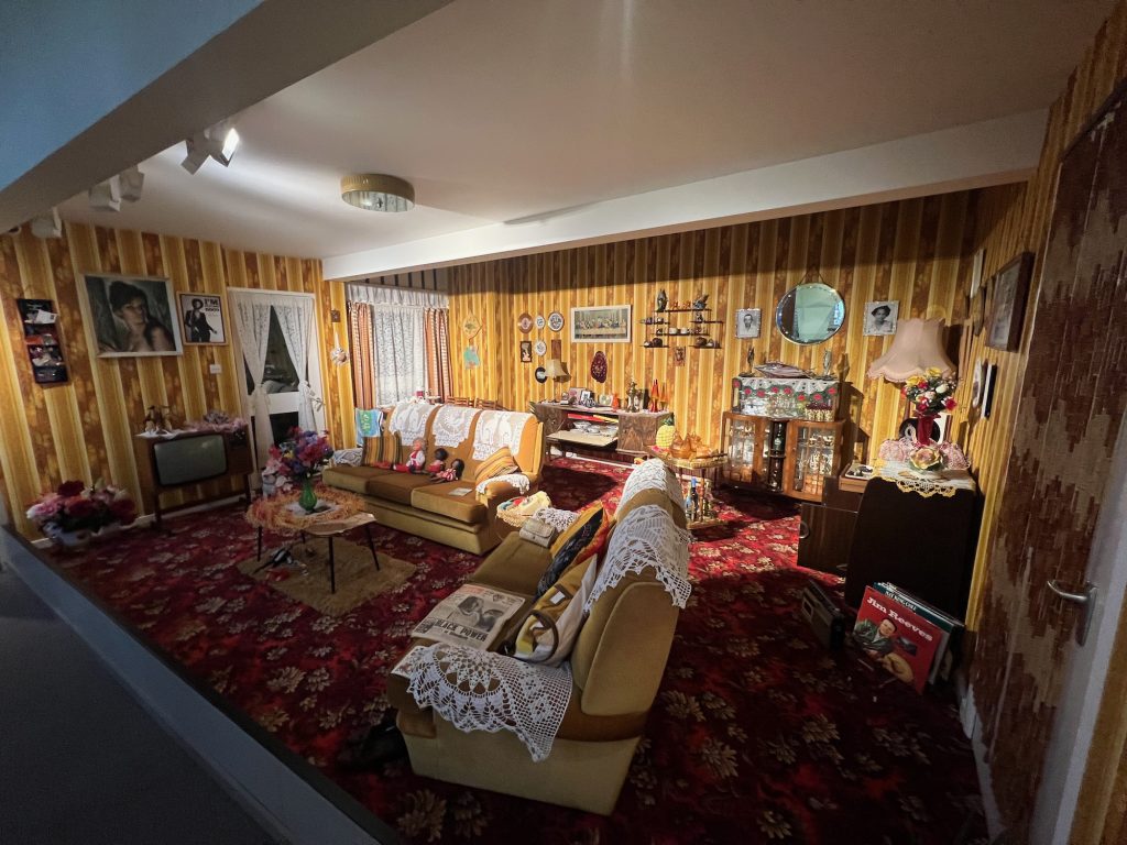 A view into a room set up almost like a stage set; three walls are decorated, and we look into the room through a large window that takes up all but the very edges of the fourth wall. The room is decorated with a striped brown patterned wallpaper. Many photographs, pictures and nick-nacks are hung from the walls, and there is lace curtain over the doors at the back left of the room. There are wooden cabinets on the back and right walls of the room, with the left wal having a TV against the wall. All are filled with items; the room looks busy but not untidy. Two brown sofas are arranged near the television, with pieces of white lace draped over the backs as headrests, and over the arms as armrests. The three on the left-hand sofa show two cats, a peacock, and another two cats. There is a bunch of pink and purple flowers in a green vase on the coffee table. The carpet is dark red, with a pattern in brighter red and light brown.