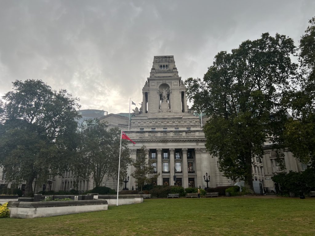 A view across a grassed square in a city, towards a large, imposting building made of Portland stone. The building is domnated by a large, stocky tower. There is a recessed archway in the front of the tower, in which there is a nude statue of a man pointing upwards and to the right. There are columns on either side of the tower, flanked by other sculptures, and there are two flagpoles either side placed on a slightly lower roof. Below, there is a grand entrance, with doors on either side of and between four Corinthian columns, each door having large windows above it on the two higher floors. Ariund the building are other Portland stone buildings, mostly masked by trees, and some modern glass buildings can be seen rising above in the background. In the foreground, on the grassed area, there is an area on the left surrounded by low falls, next to which a flagpole flies the Red Ensign. The sky is overcast and moody.