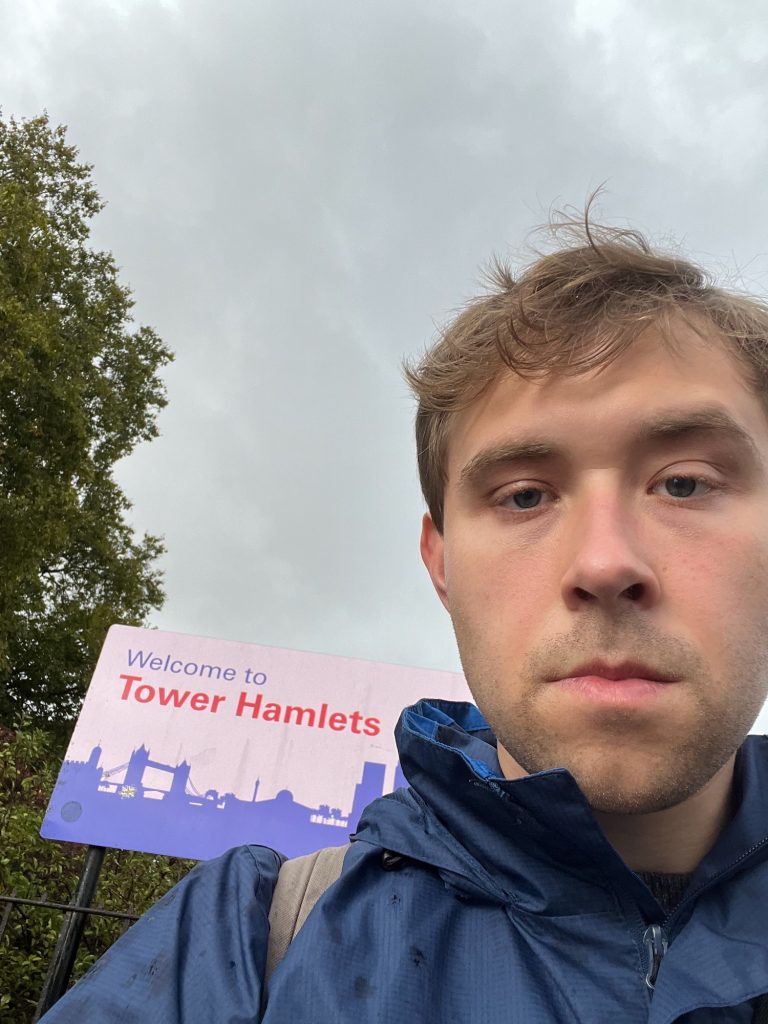 A selfie of the blog's author, who looks impassively at the camera. His hair is slightly messy; he is weirding a dark blue raincoat, and the strap of a backpack can be seen. Behind him is a sign that depicts a silhouette skyline of Tower Hamlets (including Tower Bridge and Canary Wharf); the silhouette is blue on a white background. Above that the sign says "Welcome to" in blue, and then "Tower Hamlets" in red. Behind that again, and to the left, there is a tree rising above the sign. The rest of the background is sky, which is overcast.
