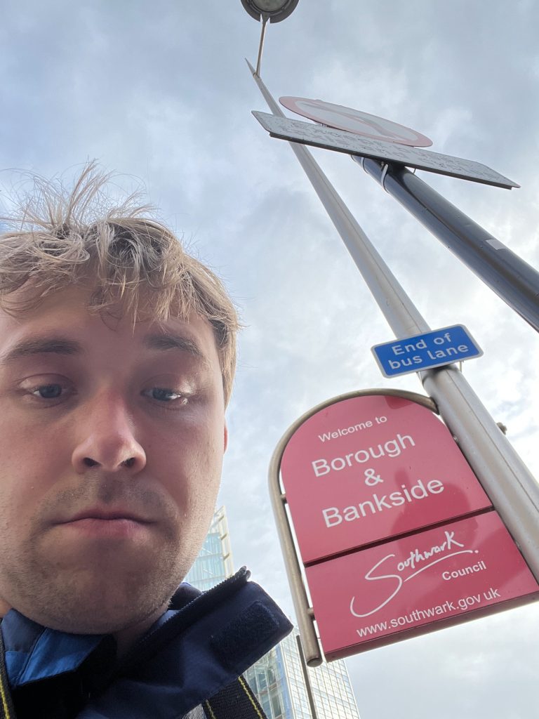 A selfie of the blog's author, looming over the camera. Behind him is a red sign in two panels, which is attached to a lamppost. The sign has an arched top, but the sides are then vertical and the bottom horizontal. The upper part of the sign says, in white text, "Welcome to BOROUGH & BANKSIDE". The lower panel, which is also white-on-red, shows the Southwark Council logo; this has the word "Southwark" as if handwritten, with a very large S that descends well below the other letters. Those other letters are underlined with a curved line, which ends with a separate dot. Below and to the right, the word "Council" is printed in a sans-serif font. Below the logo is the Southwark council web address. The sign is surrounded by a curved tube of metal following the outline of the sign from the bottom left, over the arch, and to the top-right corner, where it stops at the lamppost. The lamppost also has a small white-on-blue "End of bus lane" sign on it, and the circular downward-facing light can just be seen at the top of the lamppost. Next to the lamppost is another signpost, with the "No left turn" sign and a supplementary plaque that says "Except buses, taxis and cycles. Behind all of this can be seen some small parts of glass-walled high-rise buildings, and a cloudy sky.