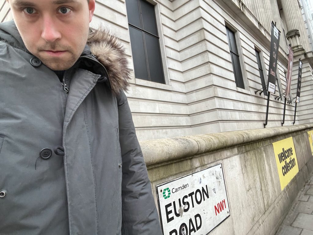 A selfie of the blog's author, who is wearing a green-grey winter coat with a faux-fur-lined hood. He stands in front of a Portland stone building; on the ground floor, there are deep horizontal grooves between successive rows of stones, and four signs angled out from the wall advertising exhibitions. Above the ground floor, it is just possible to see some classical-style columns as part of the wall above. In front of the building is a low wall abutting the pavement. On that wall, immediately to the right of the person, is a white street sign. It has the Camden council logo at the top (eight green hands, clasped in pairs, arranged in a circle, beside the word "Camden" in black). Below, in black, it says "Euston Road", and then, in red, it says "NW1". Further along the wall, to the right, is a yellow sign with "wellcome collection" written on it in yellow.