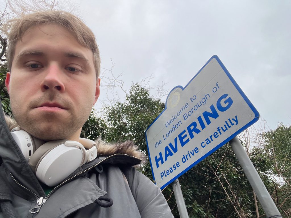 The blog's author, now wearing white Sennheiser headphones around his neck. He stands in front of a white sign with a blue border, held up by two grey poles. The sign is rectangular, except that there is a semicircle protruding from the middle of the top edge. Inside the semicircle is a faded image of a coat of arms. Below that, the sign says, in blue: "Welcome to the London Borough of HAVERING. Please drive carefully". Behind the sign is foliage, extending a little higher than the sign itself. The sky is grey.