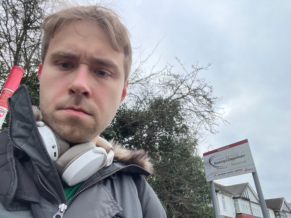A selfie of the blog's author, standing in front of foliage and a red-and-white pole marking the location of a gas main. Behind him, next to some foliage along the edge of the road, there is a white rectangular sign held up by two grey poles; it is quite a long way away, so appears small. There is a red strip along the top of the sign that says "Welcome to" on it in white. Below that, the sign says, in various shades of red, "London Borough of Barking & Dagenham", followed by the council's web address. There are two thin crescent shapes arcing around the name of the borough on the left. There is then a horizontal line across the sign, and writing below which is mostly hard to read. What can be made out is a logo made up of a red square and three grey triangles, the space between them forming an approximate "M" shape; and text reading "Myrene Signs Limited". Behind the sign, between its poles, there are some houses with mock-Tudor frontages. The sky is grey.