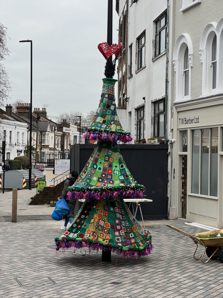 A lamppost on an urban street; there is a shop to the right, and there are houses behind. The lamppost is on a pavement made up of small stones. Around the lamppost are three cone-shaped pieces of fabric, one above the other, presumably held up by supports underneath. The fabric is made of lots of little squares sewn together, which are crocheted. Lots of colours are used in the crochet, but the predominant one is green. Around the bottom of each cone are pink and purple tassels. Above the uppermost (and smallest) cone is a tightly-wound string of fairy lights, which are off, and above that three red hearts are arranged in a ring around the lamppost.