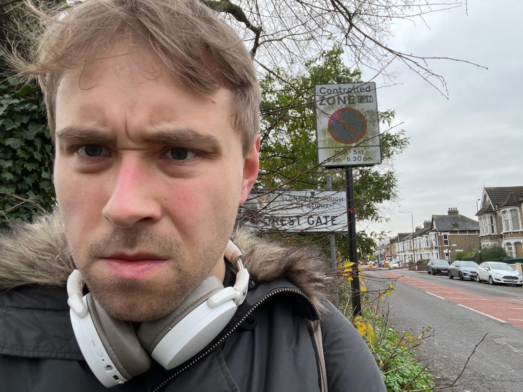 A selfie of the blog's author, standing on the edge of a road. Across the road are Victorian yellow-brick terraced houses. On this sign, behind the person is a sign for a controlled parking zone. Behind that, there is a white rectangular sign with a black border, on grey poles. It is partly hidden by the branches of a bush. It shows a coat of arms, to the right of which is written "London Borough of Newham" in black. Below that, in black, it says "Forest Gate".