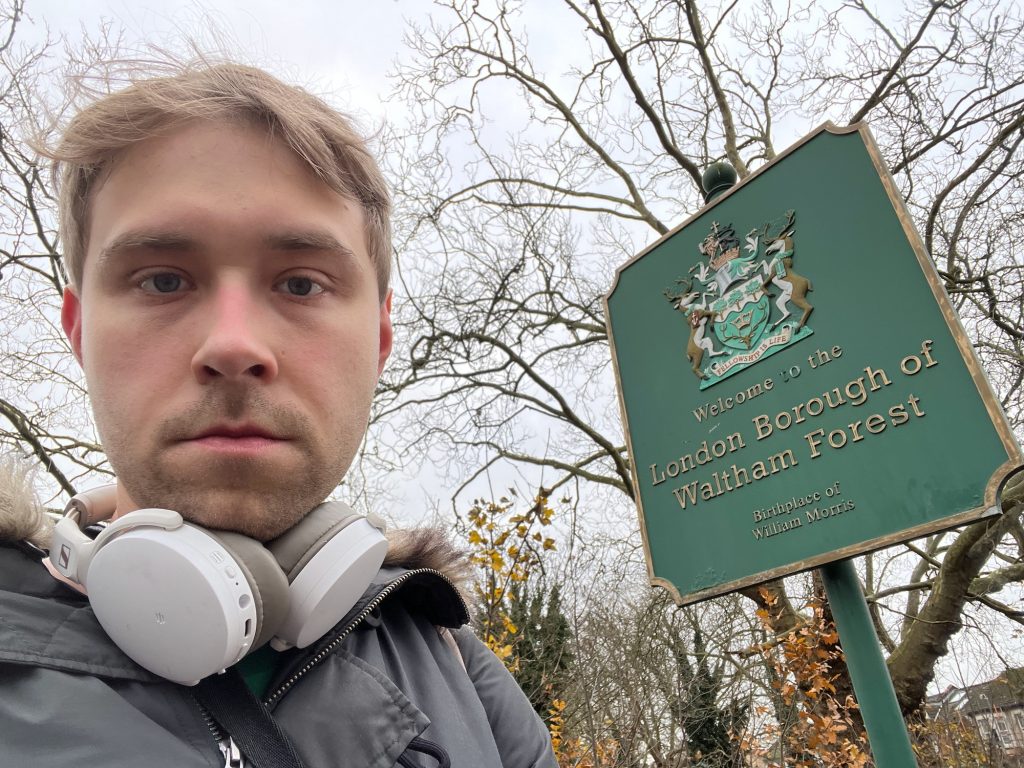 A selfie of the blog's author, standing next to a green sign on a green pole. The pole is topped by a decorative ball. The sign is rectangular, with a quarter-circle cut out from each corner, and has a raised border painted gold. The top half of the sign shows a coat of arms in bas-relief, painted in the appropriate colours. The shield is divided by a wavy line into white and green parts; on the white part at the top are three trees, and below that on the green part is the head of a stag with a crown between its horns. The supporters are two stags, and the motto is "Fellowship is Life". On the sign, below the coat of arms, are words embossed and painted gold: "Welcome to the LONDON BOROUGH OF WALTHAM FOREST, Birthplace of William Morris". Behind th person and the sign are some bare trees and other foliage.