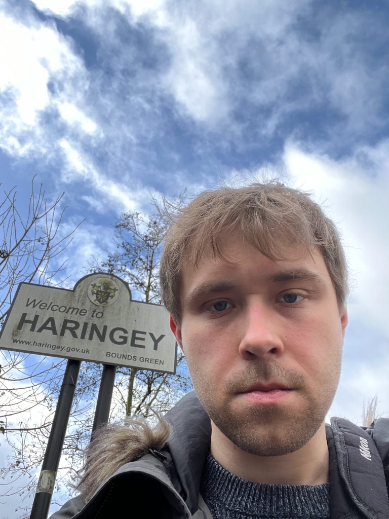 A selfie of the blog's author, wearing a thick coat and with a camera bag over his shoulder, standing in front of a sign. The sign is supported by two closely-spaced black poles. The sign itself is a rectangle, with a semicircular "bump" protruding from the midle of its upper edge; it is white, with a black border. Within the bump is a coat of arms, on a circular sticker with a white background. Below that, in black text, the sign says "Welcome to HARINGEY". Underneath again is the borough's web address, bottom-left, and "Bounds Green", bottom-right. Behind the sign are some trees with few-to-no leaves. The sky is mottled blue and white.