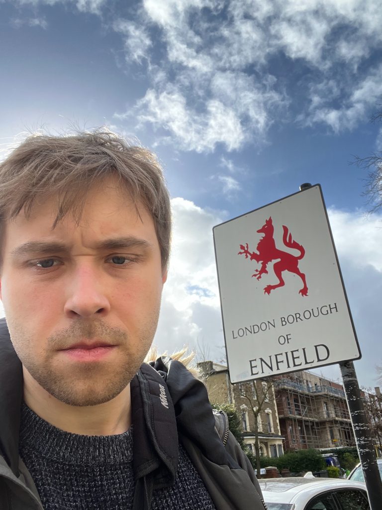 A selfie of the blog's author, next to a sign. It is a portrait-orientated rectangle with rounded corners, placed off-centre on a black pole (the pole is at the right-hand side of the sign). The sign is white with a black border. A large silhouette image (in red) of a heraldic beast takes up most of the sign; it is known as an "enfield", and has a fox's head, an eagle's forelimbs, a greyhound's chest, a lion's body, and a wolf's tail. Beneath the image are the words "London Borough of ENFIELD", in black. Behind the sign is a street with cars parked along the near side, and property visible on the far side—a yellow-brick Victorian house, and a building partly obscured by scaffolding. The sky is partly cloudy.