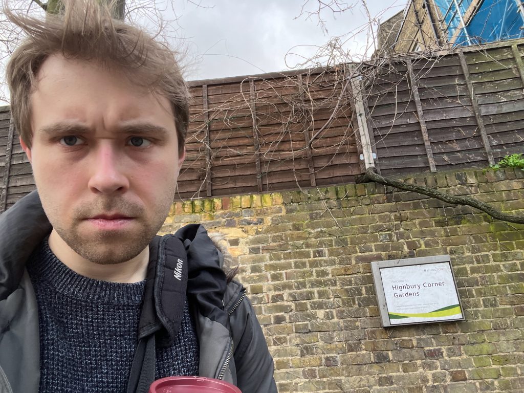A selfie of the blog's author, now holding a Pret coffee cup. He is standing in front of a brown-brick wall, which is topped by a wooden fence. On the wall is a small sign, which is barely legible, but you can make out that it says "Islington" at the top-right", and "Welcome to Highbury Corner Gardens" below. There are branches growing up the wall and the fence. Behind the fence at the upper parts of some buildings and a tree.