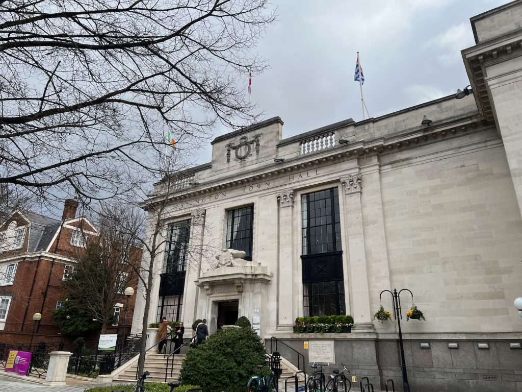 A two-storey Portland stone building. The building has an ornate portico above the front door, approached by a short flight of steps (the stonework up to near the bottom of the porch is, in fact, grey). Above the door are Roman numerals giving the date of construction as 1925. Atop the pediment over the doorway is an ornate coat of arms, carved in relief in stone. There is a large window to either side of the door, and there are three large windows on the upper floor: one is above the porch, and the others are joined by black-painted panels to the windows below. There are four shallow Corinthian columns carved into the stone to either side of the two columns of windows. The architrave above all of these has engraved text reading "Islington Town Hall". Above this is a colonnade, with a taller section above the door displaying a wreath. There are three flagpoles on the building, displaying (from left to right) the flags of Ireland, the United Kingdom, and Kiribati. The building continues to the right of the ornate frontage, but is a much plainer wall, that towards the edge of the image steps out towards the camera. There is a dedication plaque beneath the right-hand window. Several people are gathered on the top step in front of the porch. There is a sign identifying the building to the left of the steps. A three-storey red-brick building is next along the street to the left. The bare branches of a tree overhang the scene.