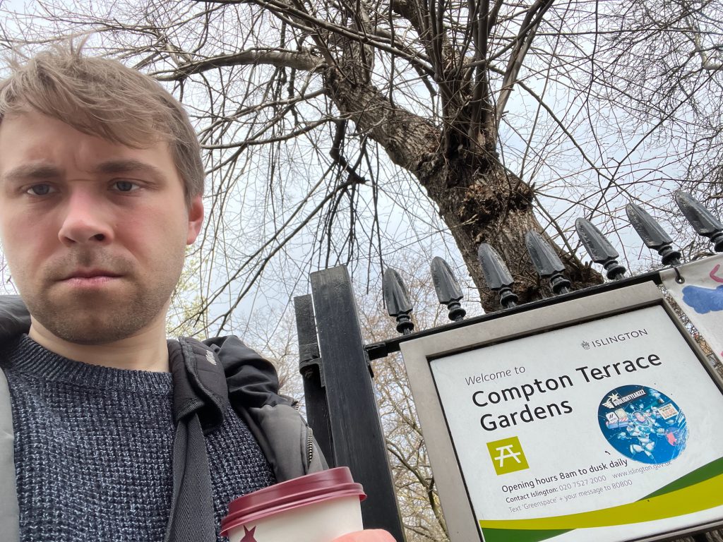 A selfie of the blog's author, still holding a coffee cup. He is standing next to a black-painted railing, to which is attached a sign, with a plain metal frame that is wide on the upper and left-hand edges and narrow on the other two. The rectangular sign inside the frame is white, with black text. At the top-right is says "Islington", to the right of a small outline drawing in black of the council's coat of arms. Slightly below that, to the left, it says "Welcome to COMPTON TERRACE GARDENS". Beneath this is an icon for a picnic table, shown in white on a lime-green square. Underneath, the sign reads "Opening hours 8am to dusk daily", and then gives contact details fo rthe council. There is a decorative lime-green "swoosh" across the bottom of the sign, with two other swooshes in darker green appearing to disappear behind it. Someone has attached a large, circular sticker with a picture of Buzz Lightyear on it to the sign. Behind the railing is a leafless tree, with a grey sky above.