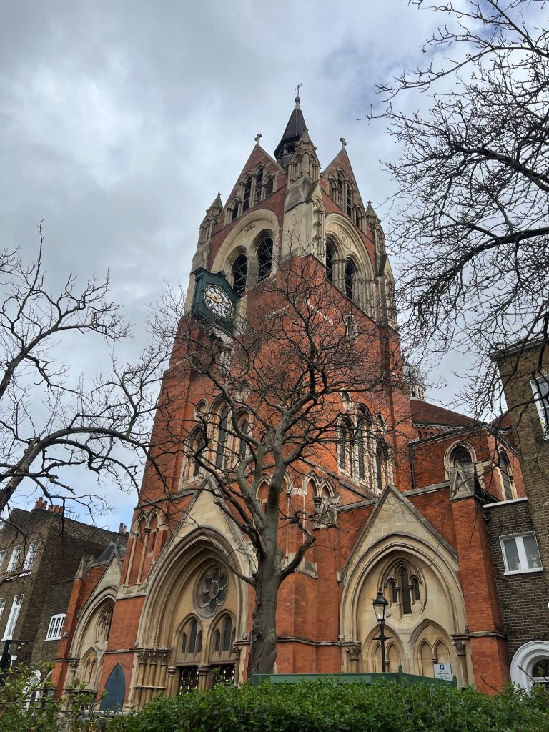 A Gothic church building, built of red brick and yellow stone. There are three recessed gothic arches, at ground floor level, with further arches recessed inside them and a pediment above each; these are about two storeys high. The rest of the fascia of the building is then dominated by a broad central tower. In the next section above the main frontage are some small windows; there is then a gothic arch on each face of the tower above that, each arch containing two arches windows. A triangular roofline tops each face of the tower, topped with a cross, and there is a spire with another cross on it behind those. There is a clock face below the big arch near the top of the tower on the front face; the clock is perpendicular to the wall of the tower. On either side of the building a houses built of brown brick. In the foreground are some trees without leaves, and a hedge. The sky is mostly grey.