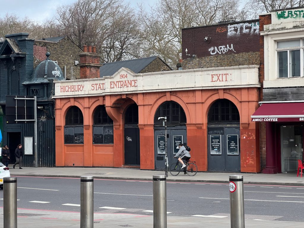 A low building whose frontage consists of five terracotta arches, above which is a white-painted architrave. The arches are rounded, with a prominent keystone; the two on the left are walled up to about a metre (with windows above), and the others contain doors, although these are firmly sealed. Above each of the three arches on the left is a word on the architrave: "Highbury", "Station", and "Entrance". (Above the word "Entrance" is the date 1904, within a triangular pointed part of the roofline.) Above the rightmost arch, again on the architrave, it says "Exit". The doors in the two right-hand arches have black posters displayed on them, with white writing. A variety of other, slightly taller, buildings, surround the station on three sides. The frontages faces a road, where someone cycles past the station on the pavement outside it. On the near side of the road are four metal bollards.