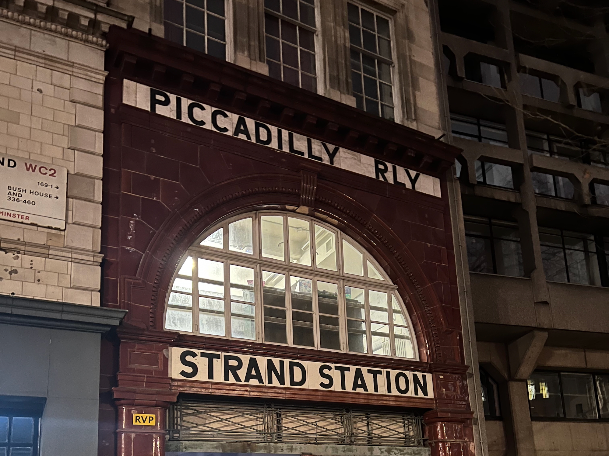 The front of a Tube station building at night. Only the first-floor level and the top of the gated portal to the ticket hall are visible. The front of the station is tiled with oxblood-coloured tiles, which surround a large arched window divided into smaller panes by white frames; the lights are on in the room behind the window, revealing some sort of office. Above and below the archway are two strips of white tiles instead of deep red, with the upper one having black letters on it that say "Piccadilly Rly", and the lower one reading "Strand Station" in similar letters. The tiled frontage is set into a larger stone building; to the left of the station, there is a blue shopfront at street level, and a street sign for the Strand on the wall. Behind the building, to the right, is a fairly tall Brutalist building.