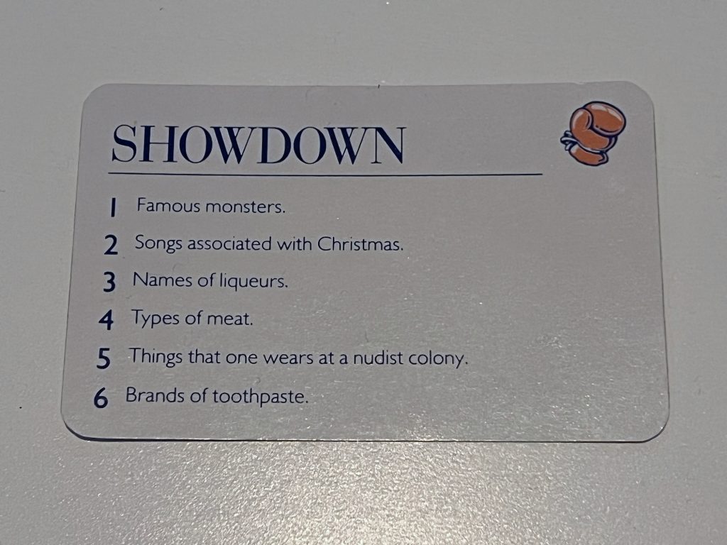 A card from a board game on a white table, entitled "Showdown". The text is blue. There is an icon of a red boxing glove at the top right. The text of the card is a list numbered from 1 to 6, with entries: "Famous monsters", "Songs associated with Christmas", "Names of liqueurs", "Types of meat", "Things that one wears at a nudist colony", and "Brands of toothpaste"