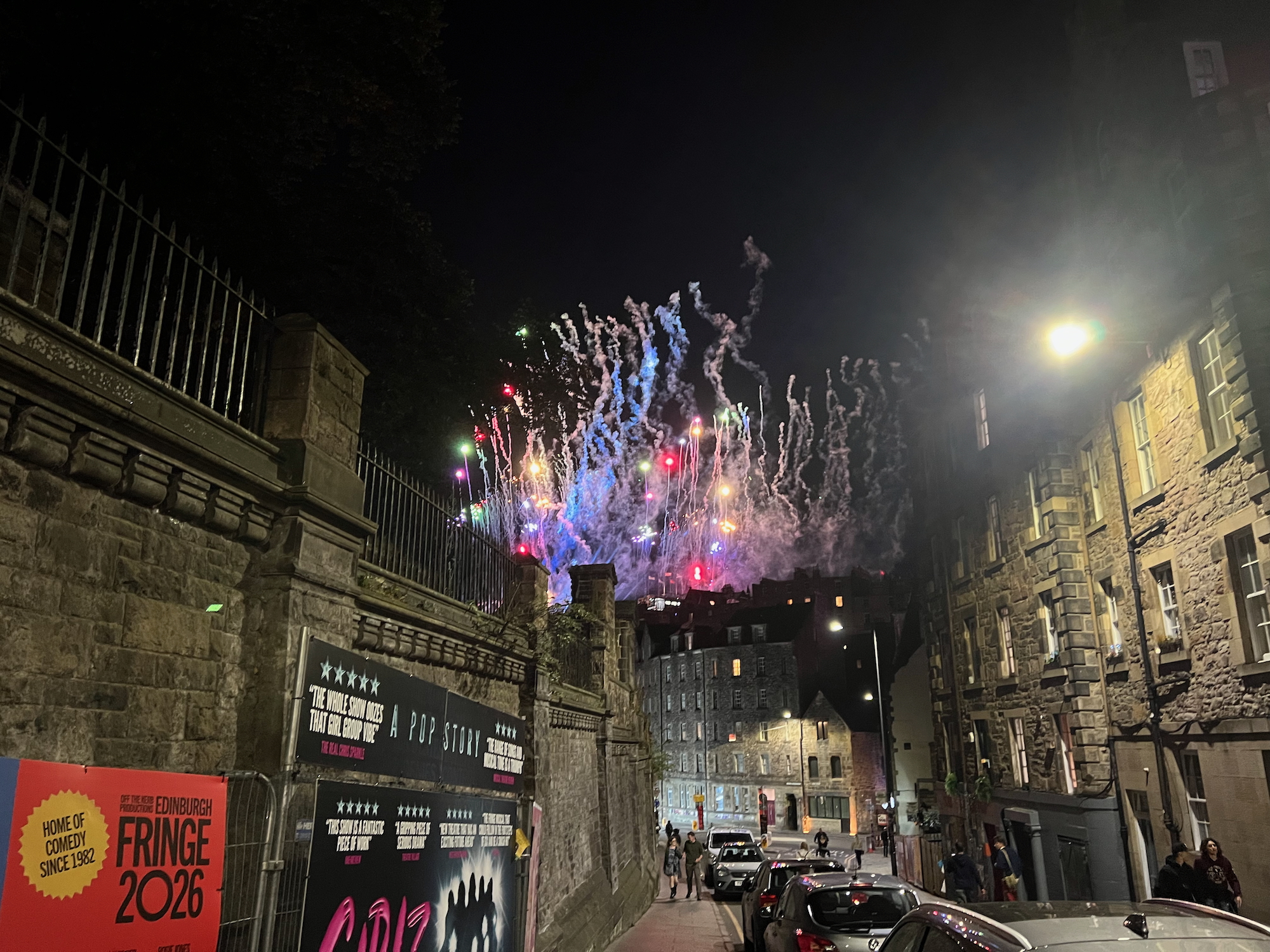 A city centre street at night. The street is on a hill, and the view is down it between a high wall on the left (topped with a railing) and some tall buildings on the right, made of stone on both sides. Cars are parked along the street, and there are some large posters near the photographer on the left. There are a few people nearby. At the bottom of the street, above a tall building, there are fireworks in the sky.