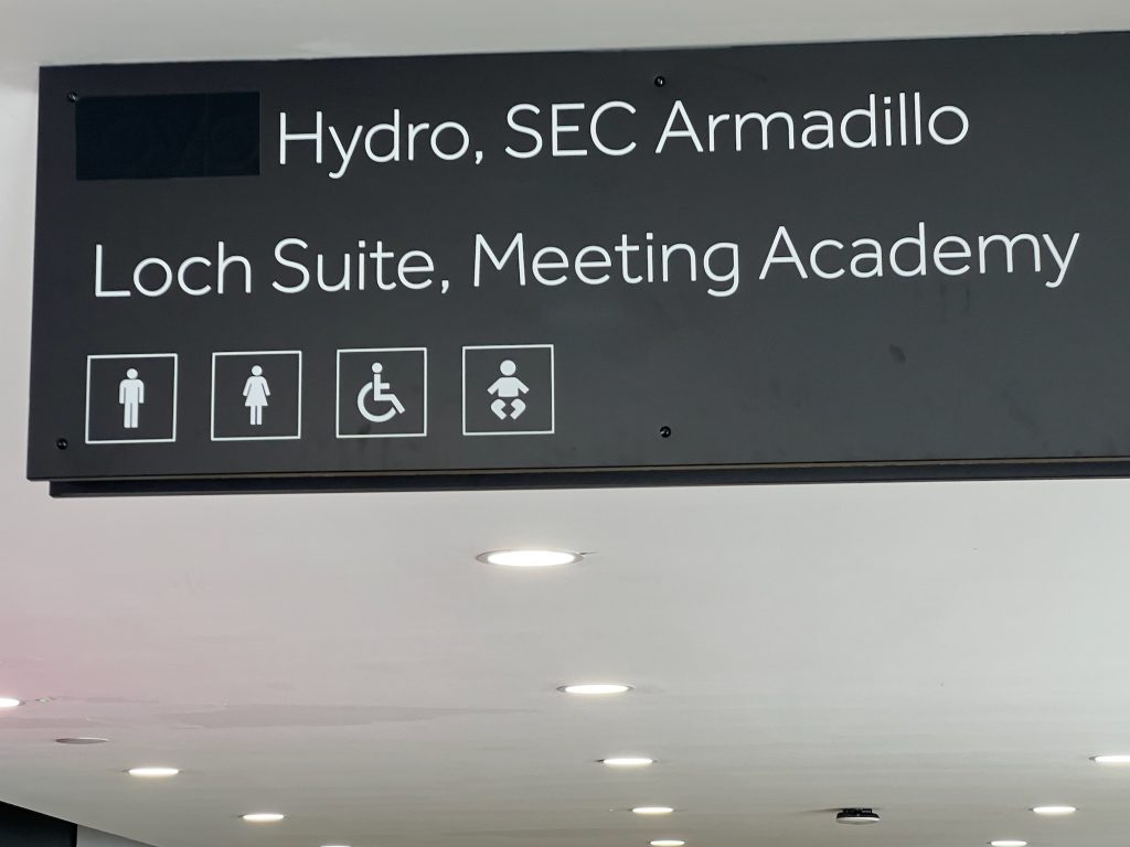 A grey sign with white writing, attached to the ceiling of a corridor. The top line of the sign has a word blacked out with a plain overlay that is a slightly darker shade of grey. After that, the text reads "Hydro, SEC Armadillo". The second line says "Loch Suite, Meeting Academy", and then there are pictograms for the men's, women's and accessible toilets, and for the baby changing room.