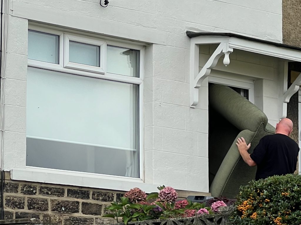 The ground floor of a house, which has a stone fascia below the window and is white render above. There is a large window, and a door beneath a porch. A sofa is being pulled through the door; a bald, white man in a black t-shirt is looking away from the camera as he tries to manoeuvre it out. A small amount of foliage appears at the bottom of the picture.