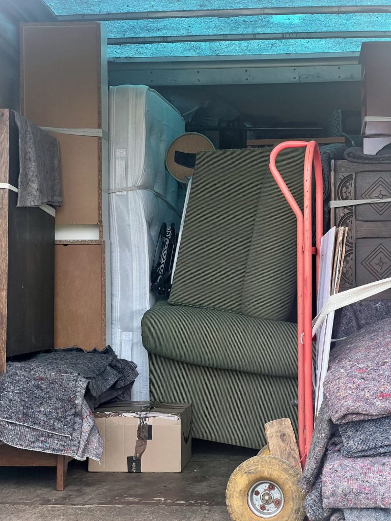 The inside of a removal van, lit mainly by its blue translucent panelled ceiling. In the middle of the van is a green corduroy sofa, on its back, perched atop another that is the right way up. Around then are other pieces of furniture, include a bed and mattress on the left, and a blanket chest on the right. There is a two-wheeled hand trolley, painted red, in the foreground.