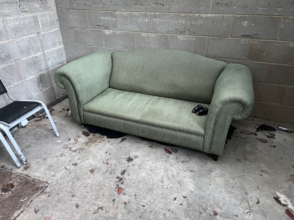 A green corduroy sofa, with two spare sofa legs resting on the seat bottom. The cushions are missing. It sits on a concrete floor in a small room with breezeblock walls. To the left, partly cut off, are two metal chairs with black seats.