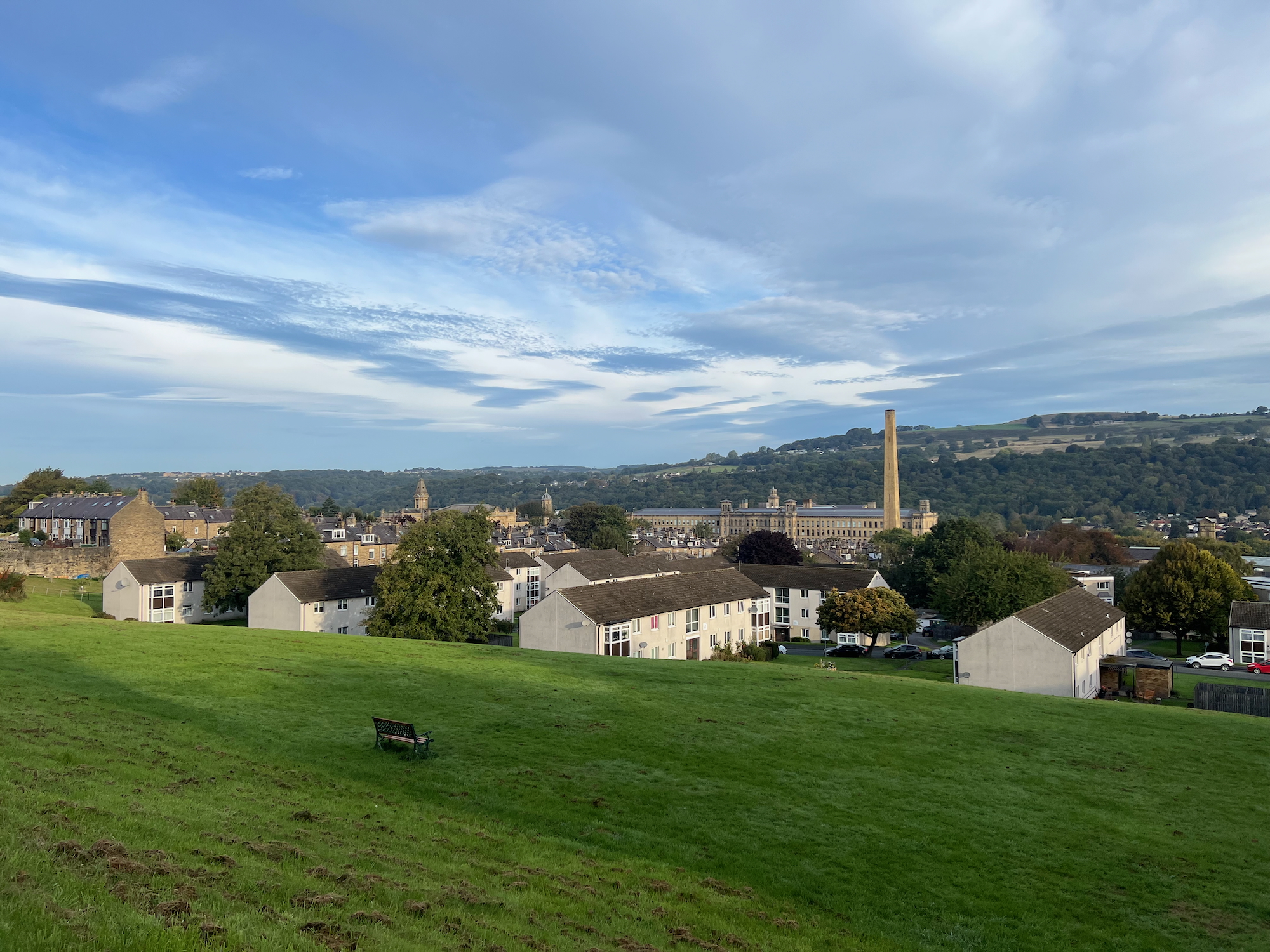 A view from the top of a grassy hill. The view is dominated by Salt's Mill, a large sandstone building with a tall chimney. To the left are some other Saltaire buildings. In front is housing, both old stone terraces and mid-20th-century blocks of flats. The grass is empty, except for a solitary bench where it flattens out a little. In the background a wooded hill towers over the scene. The morning sky is partly cloudy.